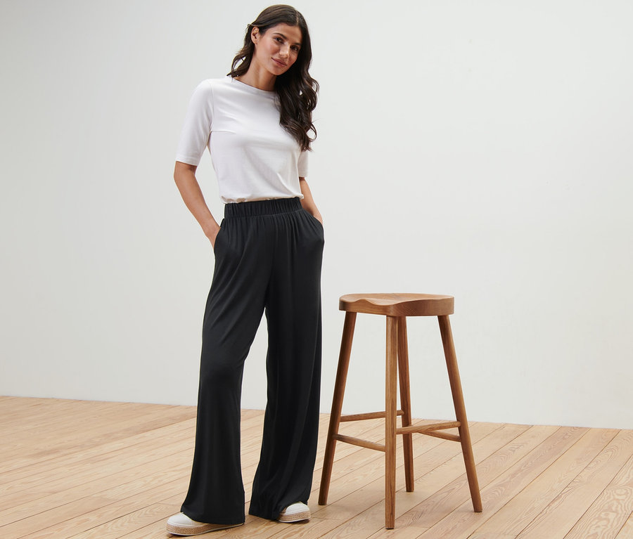 Eine Frau posiert in einer schwarzen Palazzohose neben einem Holzhocker.