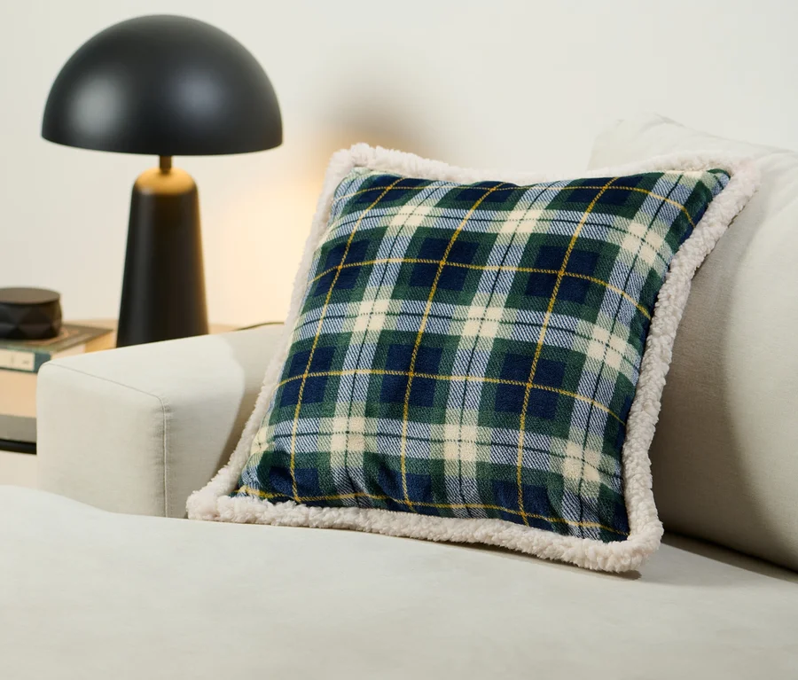 Ein kariertes Plüsch-Dekokissen liegt auf einem cremefarbenen Sofa neben einer schwarzen Lampe.