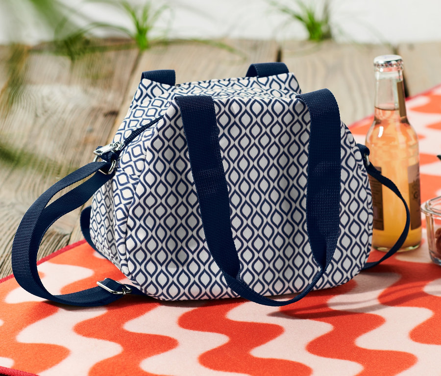 Eine blau-weiße Mini-Kühltasche liegt neben einem Getränk und einer Schüssel Pasta auf einem orange-weiß gestreiften Strandtuch.