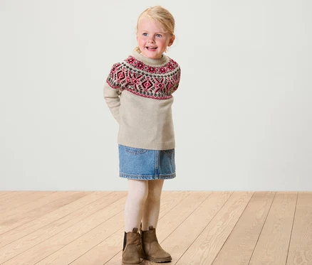 Lächelndes blondes Mädchen trägt einen Kinder-Norweger-Strickpullover, einen Jeansrock, weiße Leggings und braune Stiefel.