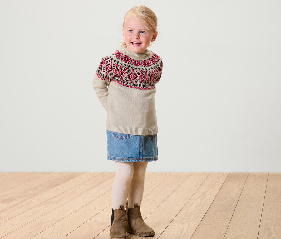 Lächelndes blondes Mädchen trägt einen Kinder-Norweger-Strickpullover, einen Jeansrock, weiße Leggings und braune Stiefel.