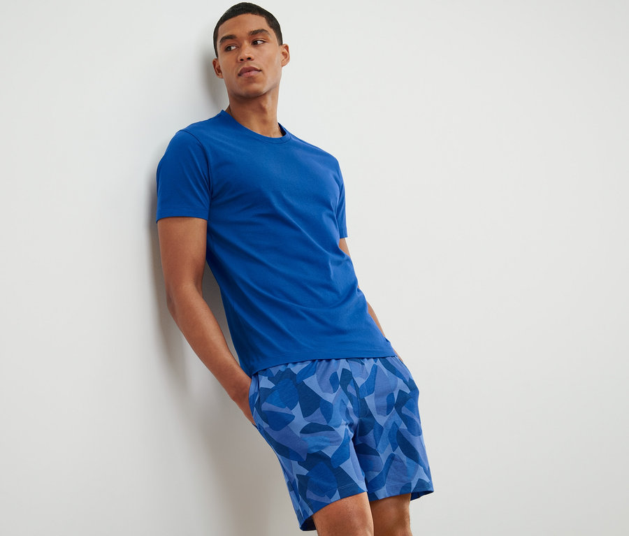 Mann lehnt an der Wand und trägt einen blauen Shorty-Pyjama.