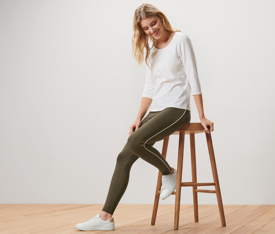 Blonde Frau sitzt auf einem Holzhocker und trägt grüne Leggings mit Paspelierung, ein weißes Shirt und Turnschuhe.