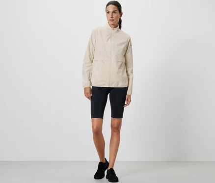 Frau steht und trägt eine creme Windprotection-Laufjacke und schwarze Sportshorts.