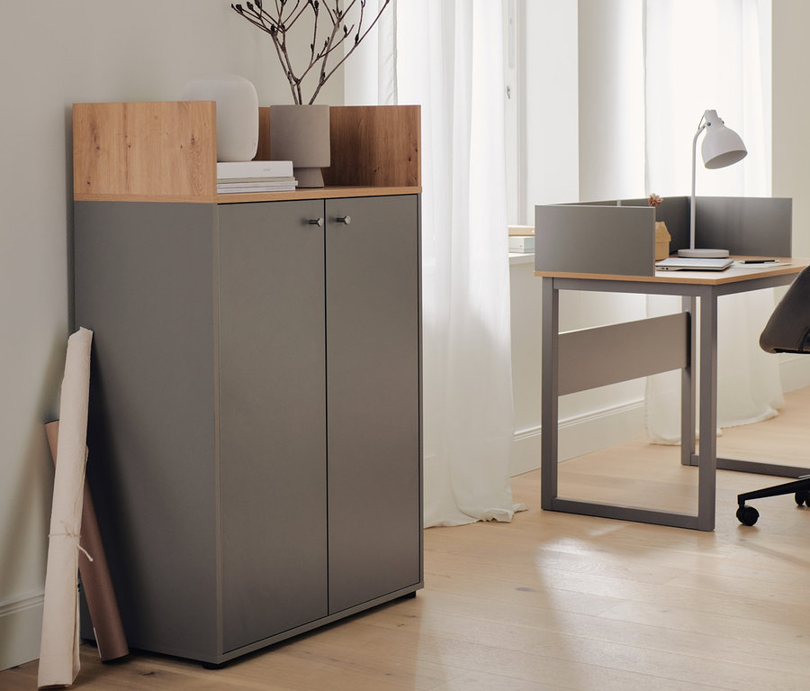 Ein grauer Sekretär mit seitlicher Umrandung und ein Highboard mit 2 Türen stehen nebeneinander. Auf dem Sekretär steht eine Tischleuchte aus Metall.