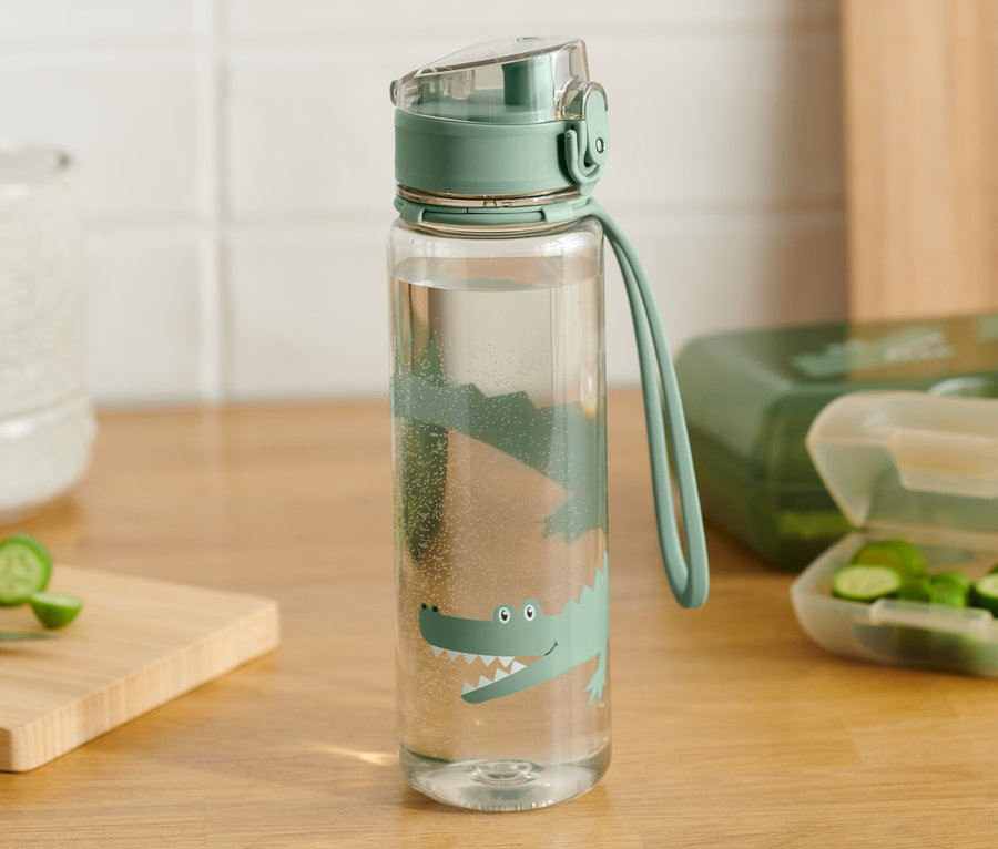 Eine beige Trinkflasche mit Krokodilprint steht auf einem Holztisch. Daneben liegen geschnittene Gurken und eine grüne Lunchbox.