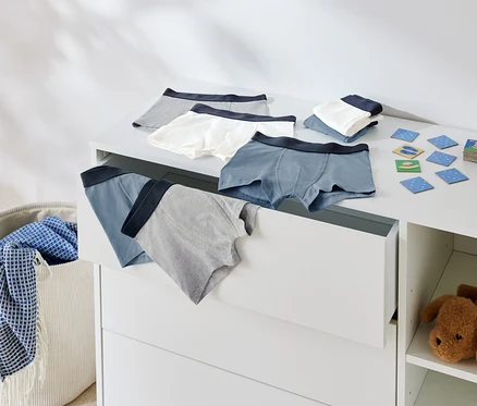 Ansicht von Kinderunterwäsche auf einer Kommode und in einer offenen Schublade, neben einem Wäschekorb und einem Stofftier.