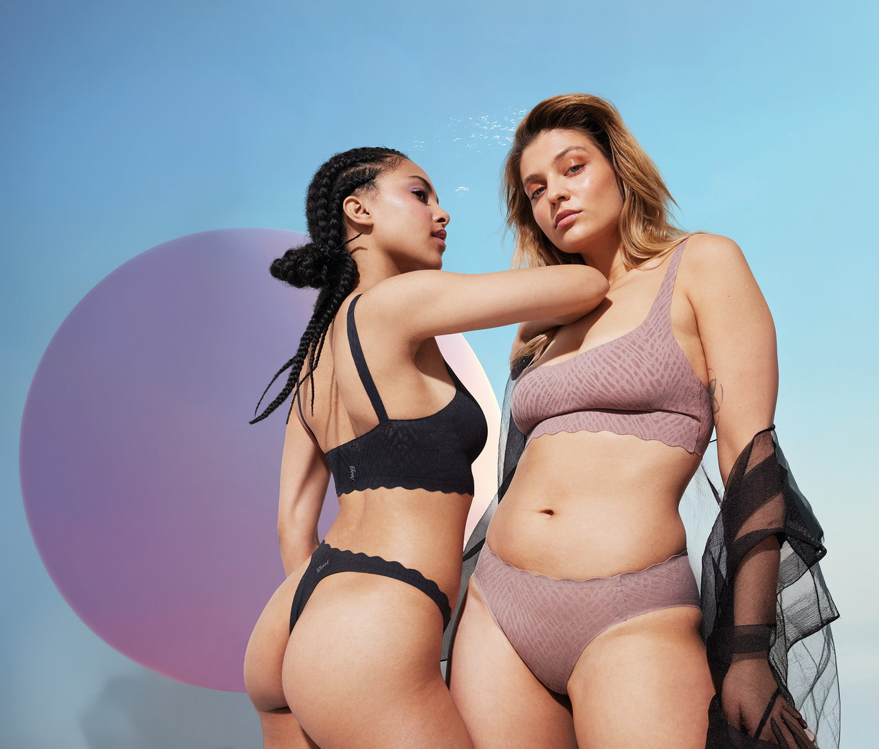 Zwei Frauen posieren, eine trägt einen schwarzen sloggi ZERO Feel Bliss Top, die andere ein gemustertes Set.