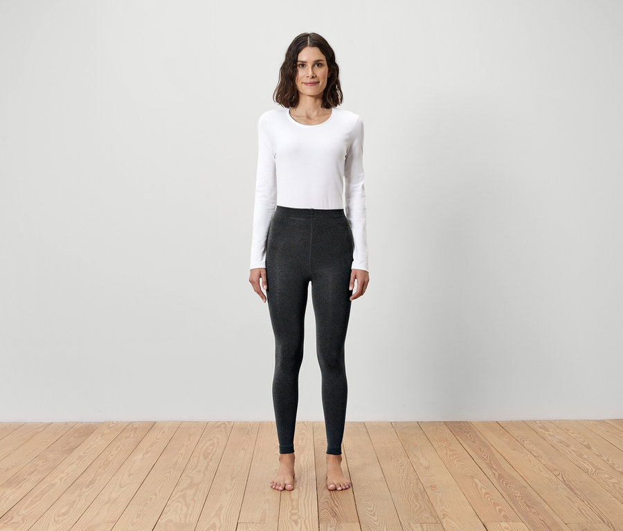 Frau steht barfuß auf Holzboden und trägt ein weißes Langarmshirt und graue Leggings.