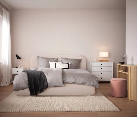 Schlafzimmer mit beigem Bett, grauer Bettwäsche und beigem Teppich. Im Hintergrund weißer Nachttisch und Kommode.
