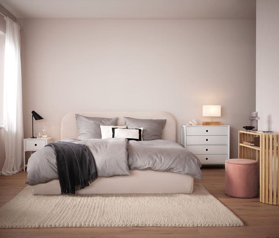 Schlafzimmer mit beigem Bett, grauer Bettwäsche und beigem Teppich. Im Hintergrund weißer Nachttisch und Kommode.