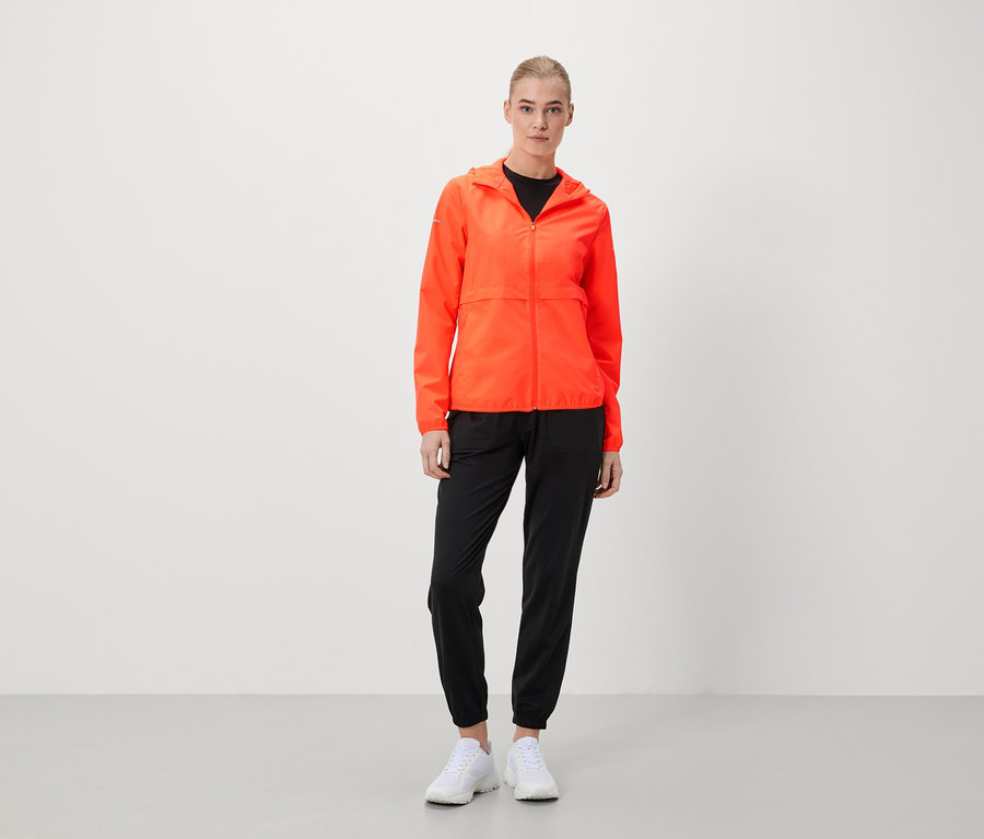 Eine Frau steht und trägt eine orangefarbene Jacke, eine schwarze Hose und weiße Schuhe.