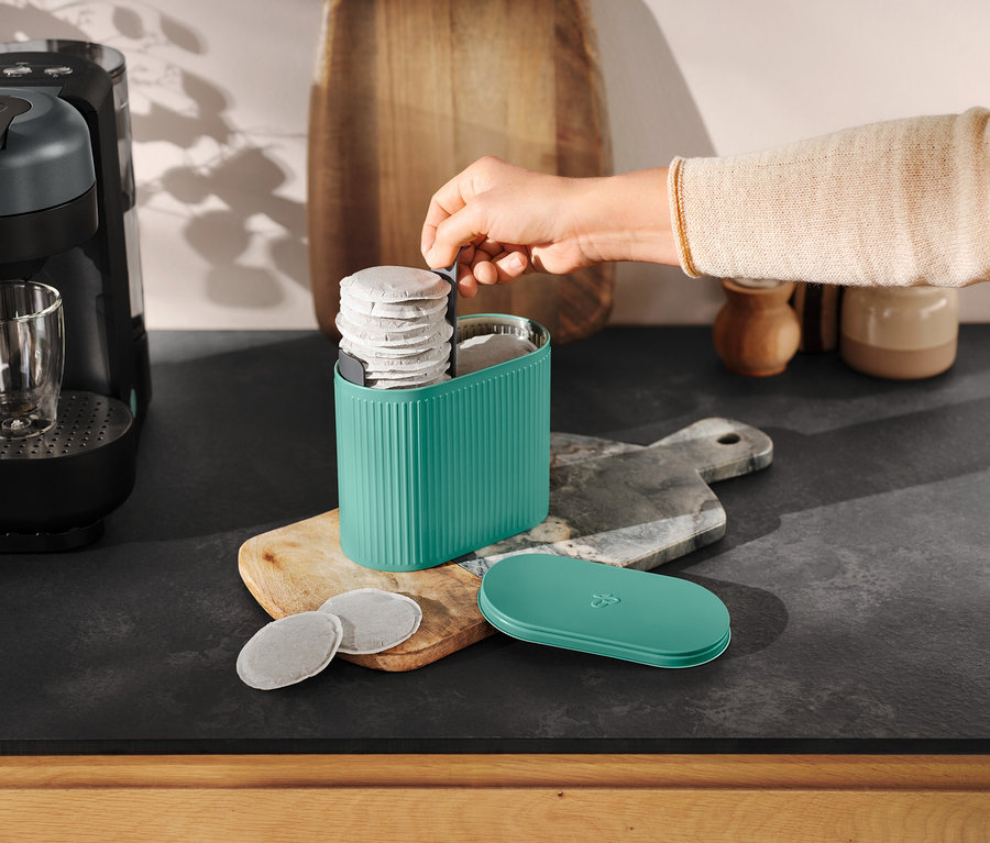 Eine Hand greift in einen türkisfarbenen Pad Box neben einer Kaffeemaschine.