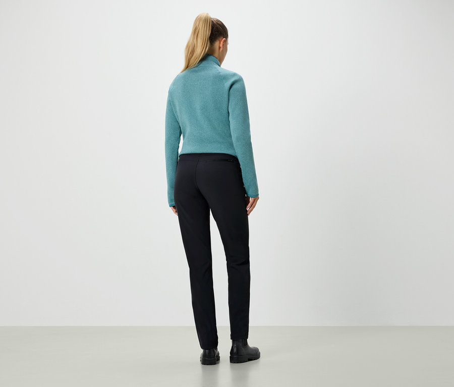 Rückansicht einer Frau mit blonden, zum Pferdeschwanz gebundenen Haaren, die einen blaugrünen Pullover und eine schwarze Hose trägt.