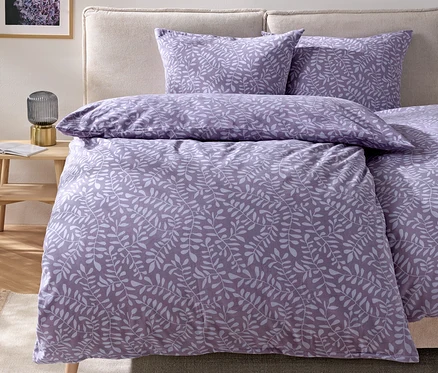 Bettwäsche mit lila Blättermotiv auf einem Bett mit beigem Kopfteil.