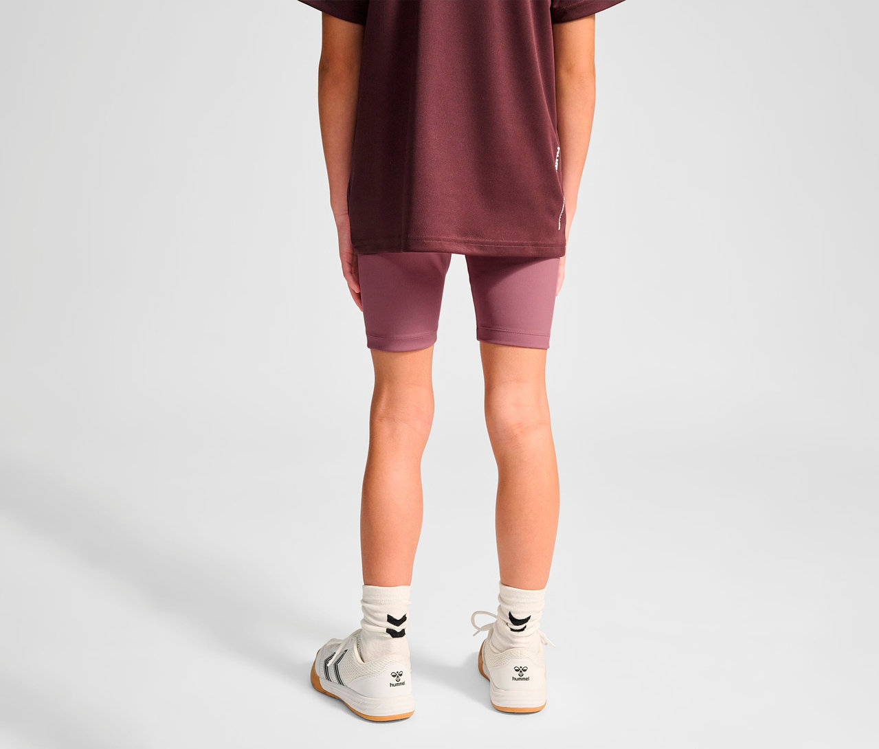 Eine junge Person von hinten trägt lila HUMMEL HMLJR Pulse MW Short Tight und weiße Turnschuhe.