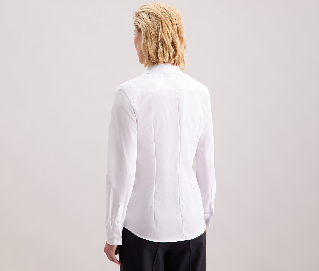 Rückansicht einer Frau mit blonden, schulterlangen Haaren, die eine weiße, schmal geschnittene, bügelfreie Seidensticker Bluse trägt.