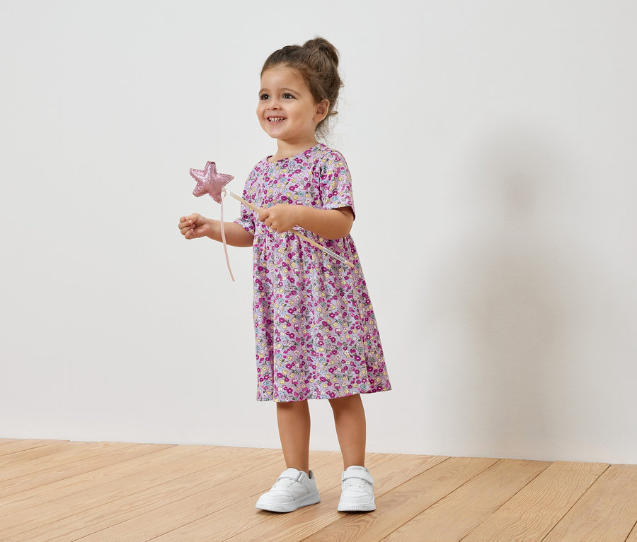 Lächelndes kleines Mädchen hält einen Sternenstab und trägt ein geblümtes Kleid und Turnschuhe.