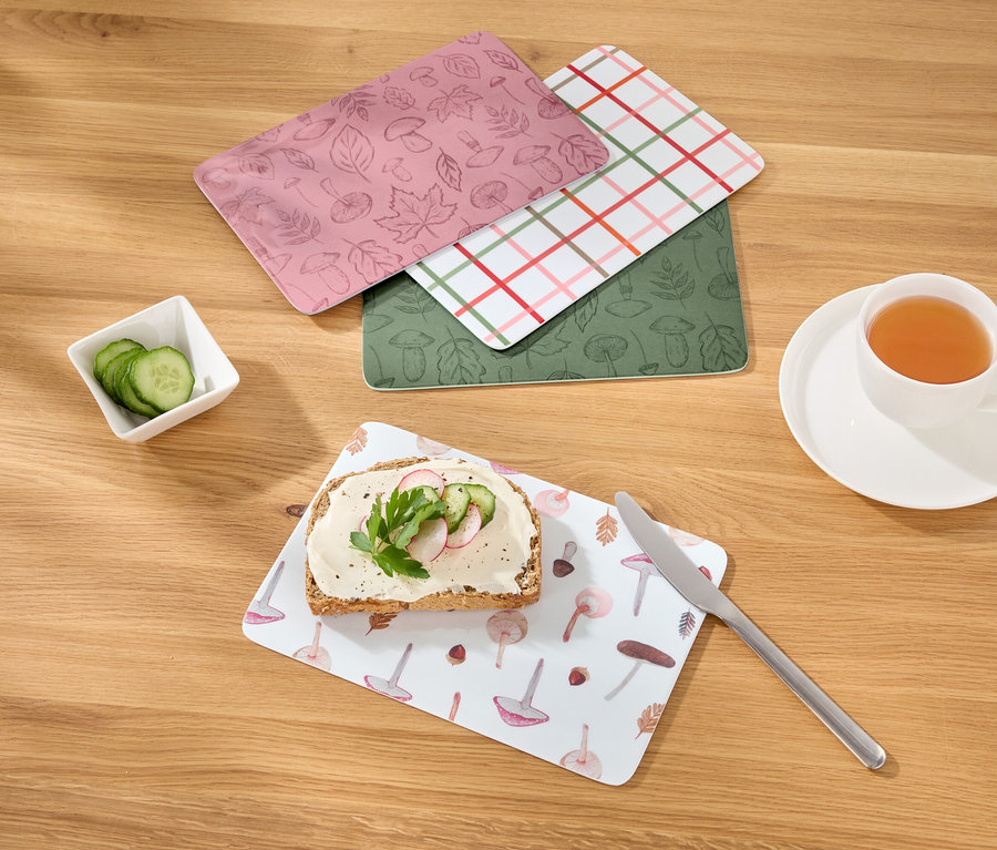 Auf einem Holztisch liegen vier Frühstücksbrettchen, ein belegtes Brot, Gurkenscheiben und eine Tasse Tee.