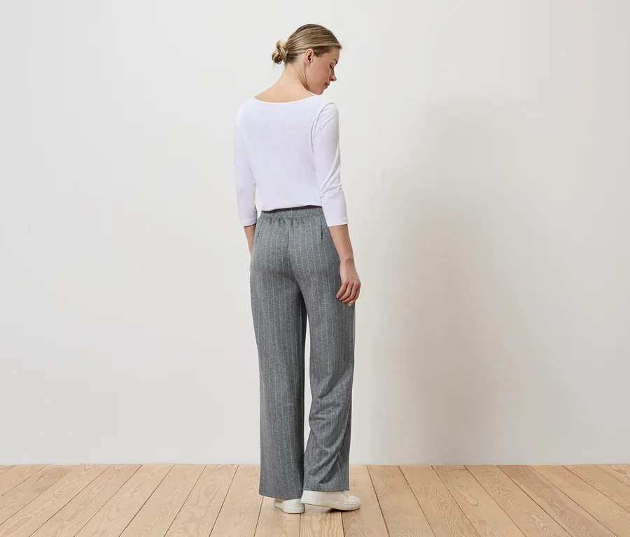 Eine Frau mit Dutt trägt ein weißes Langarmshirt, eine graue, gestreifte Hose und weiße Schuhe.