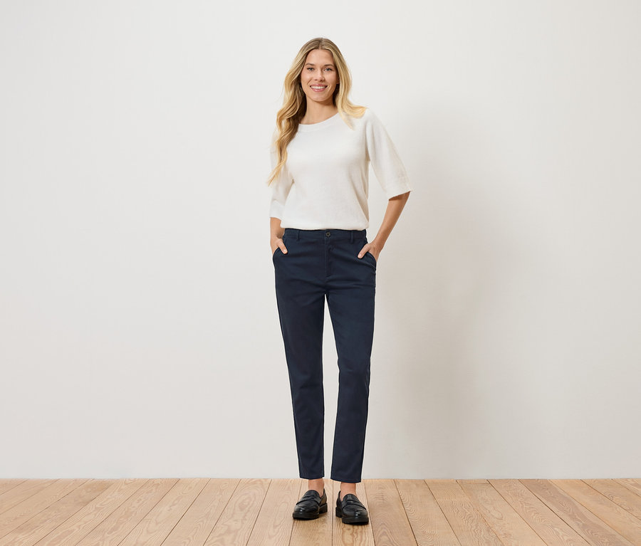 Eine Frau mit blonden, gewellten Haaren steht mit den Händen in den Hosentaschen da. Sie trägt einen weißen Pullover, eine marineblaue Hose und schwarze Schuhe.