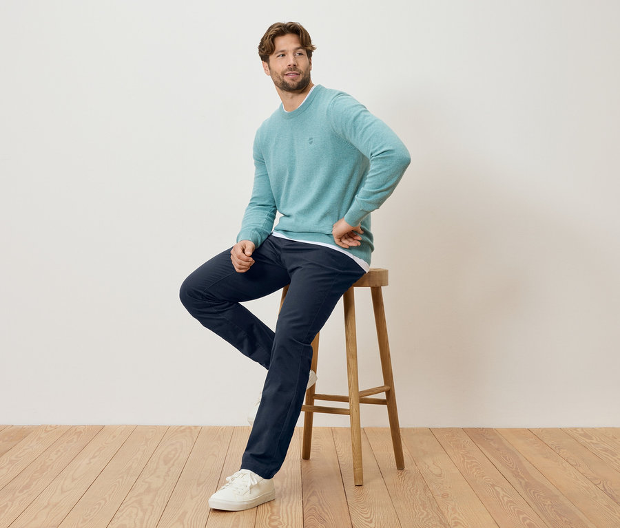 Mann sitzt auf einem Hocker, trägt einen mintfarbenen Pullover, eine marineblaue Hose und weiße Turnschuhe.