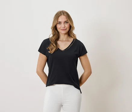 Frau mit blonden Haaren trägt ein schwarzes T-Shirt mit V-Ausschnitt und weißen Hosen vor einem neutralen Hintergrund.