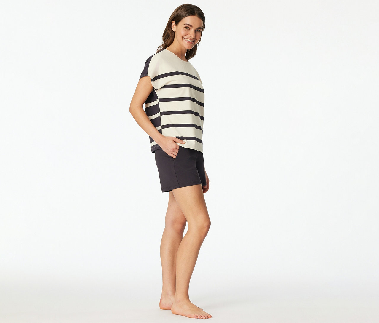 Frau trägt ein SCHIESSER Shorty-Set mit gestreiftem Oberteil und dunklen Shorts, lächelt und hat die Hand in der Tasche.