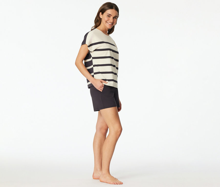 Frau trägt ein SCHIESSER Shorty-Set mit gestreiftem Oberteil und dunklen Shorts, lächelt und hat die Hand in der Tasche.