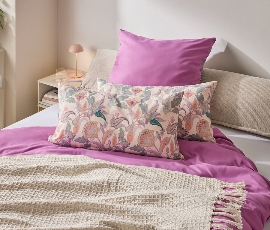 Ein Schlafzimmer mit einem Bett, das mit einem lila Bettbezug, Kissen mit Blumenmuster und einer cremefarbenen Decke mit Fransen geschmückt ist.