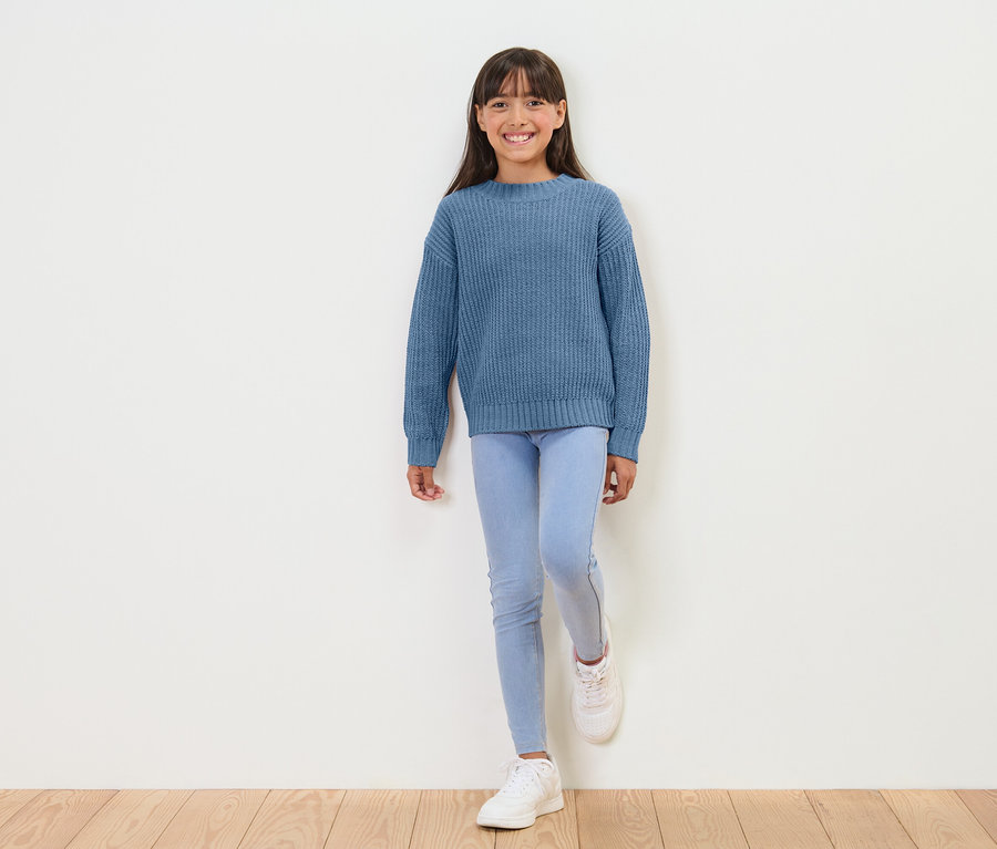 Mädchen steht vor einer weißen Wand und trägt einen blauen Pullover, hellblaue Leggings und weiße Turnschuhe.
