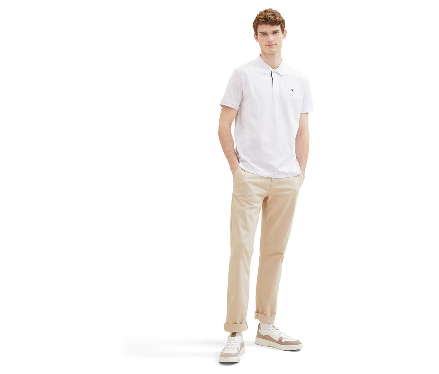 Mann in beigen Hosen und weißem Tom Tailor Piqué Poloshirt. Er steht mit Händen in den Hosentaschen vor einem weißen Hintergrund.