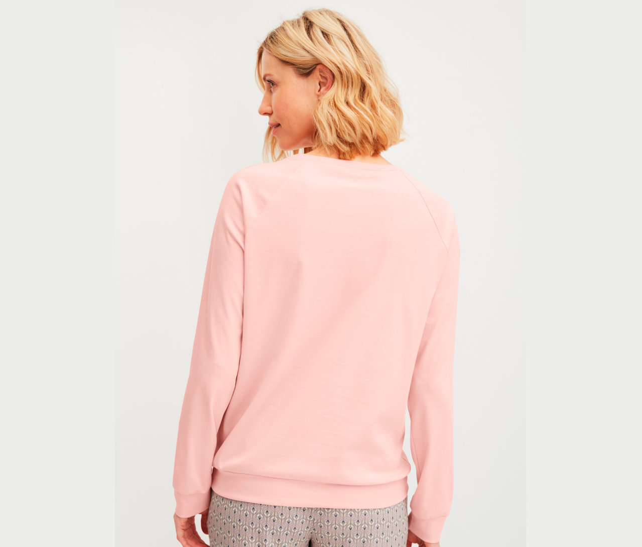 Rückansicht einer Frau mit blonden Haaren, die einen rosafarbenen Pullover und gemusterte Hosen trägt.