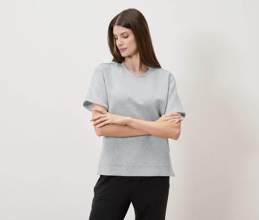 Frau mit verschränkten Armen trägt ein graues T-Shirt und eine schwarze Hose vor einem neutralen Hintergrund.
