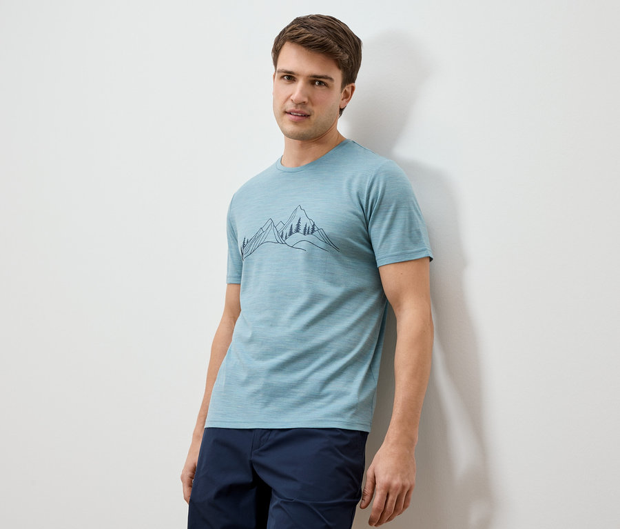 Ein Mann lehnt an einer Wand und trägt ein hellblaues T-Shirt mit einer Berglandschaftszeichnung und dunkelblaue Shorts.