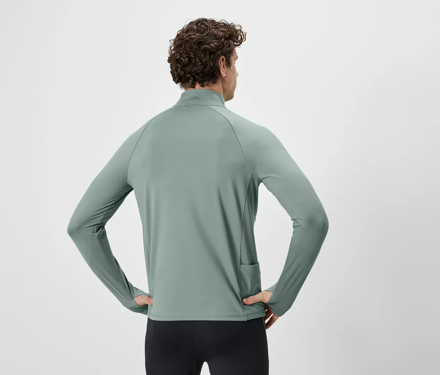 Mann von hinten, Hände in den Hüften, trägt ein grünes Langarmshirt mit Reißverschluss und Daumenlöchern, sowie eine schwarze Hose.