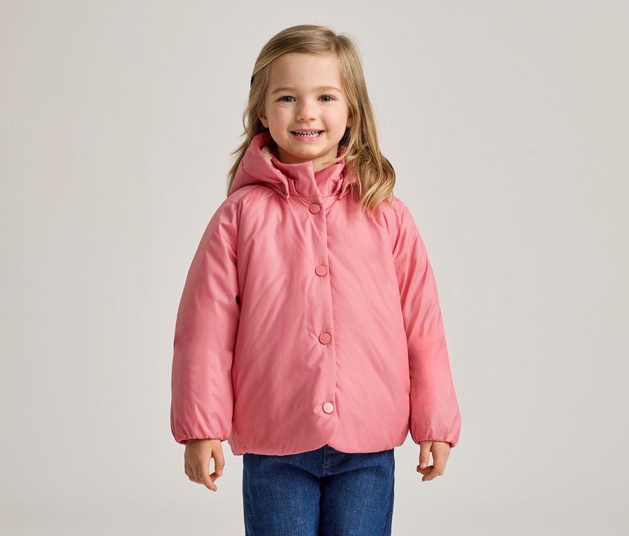 Ein junges Mädchen mit blonden Haaren, das eine rosa Kapuzen-Steppjacke und blaue Jeans trägt, lächelt in die Kamera.