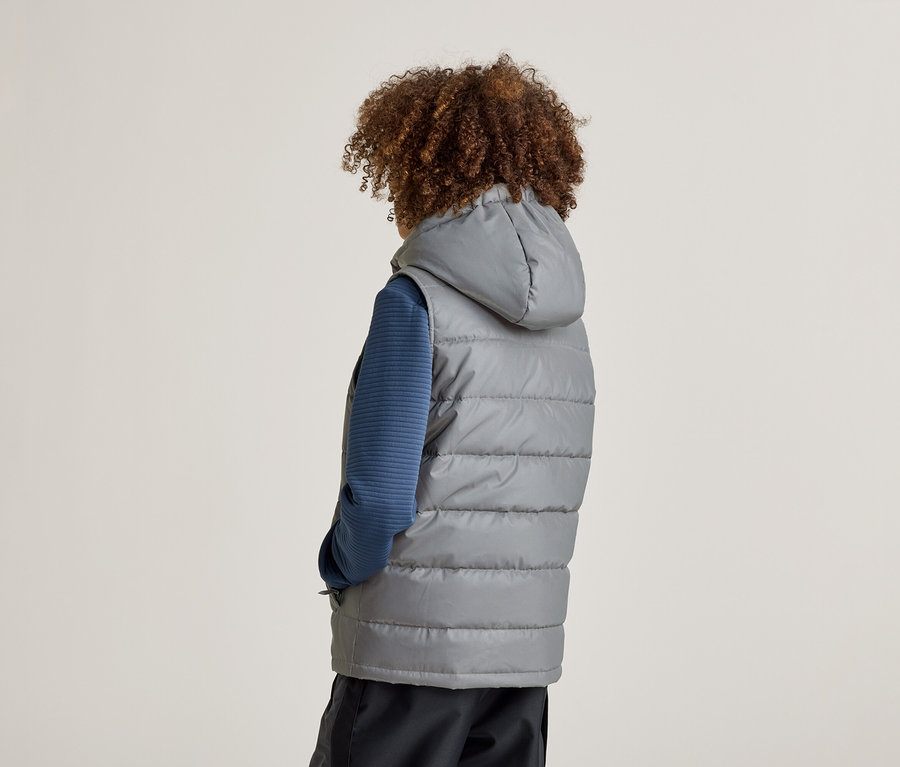 Rückansicht einer Person mit lockigem Haar, die eine graue, gesteppte Weste mit Kapuze über einem blauen Oberteil und einer dunklen Hose trägt.