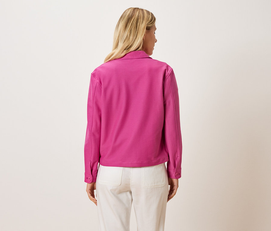 Rückansicht einer Frau mit blonden Haaren, die eine pinke Jacke und helle Hose trägt.