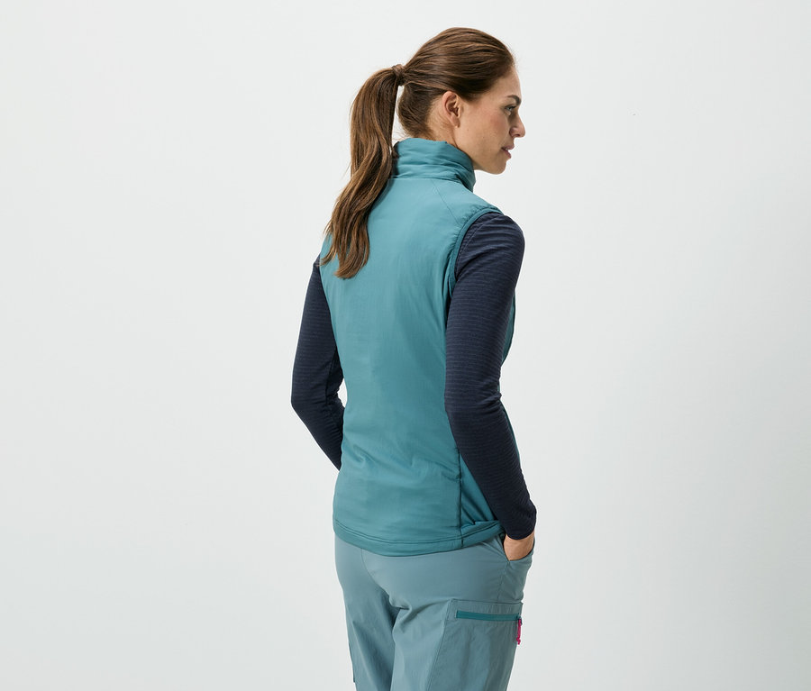 Rückansicht einer Frau mit Pferdeschwanz, die eine ärmellose, türkisfarbene Weste, ein blaues, langärmeliges Hemd und eine türkisfarbene Hose trägt und ihre Hände in den Taschen hat.