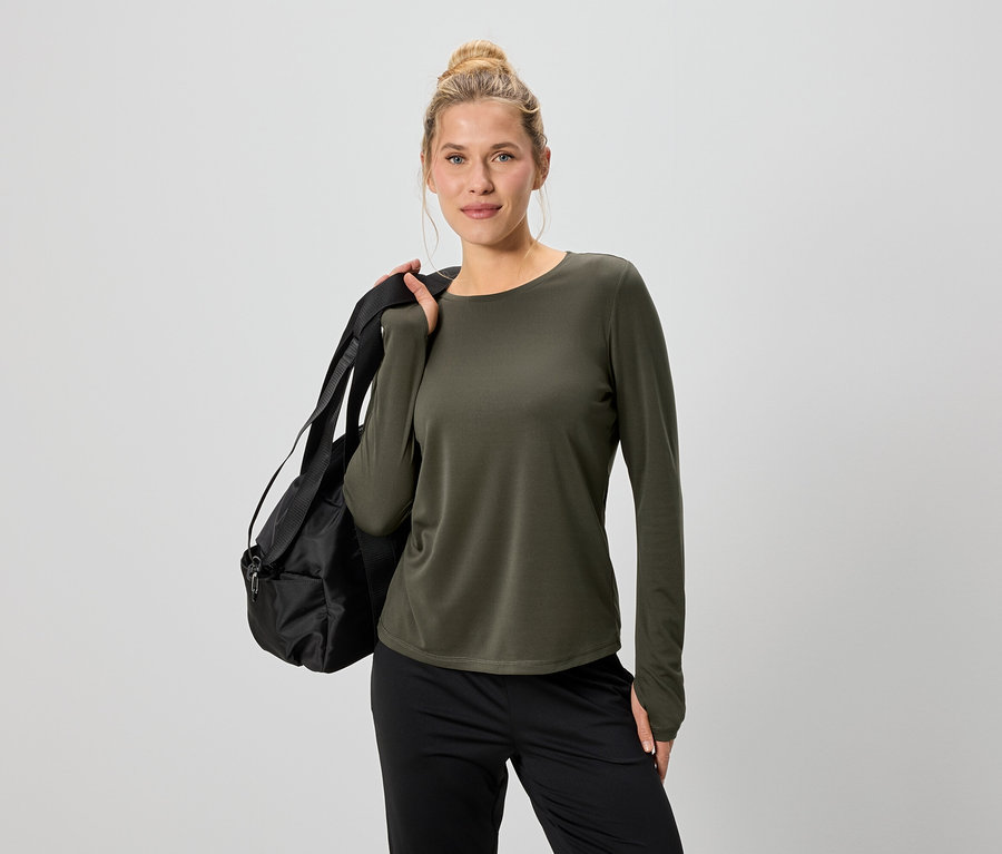 Frau mit Dutt trägt ein olivgrünes Langarmshirt und schwarze Hose, mit einer schwarzen Sporttasche über der Schulter.