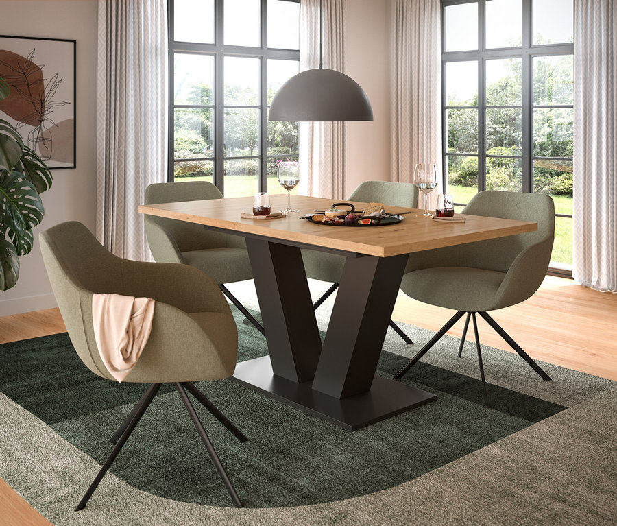 Ein Esszimmer mit einem Holztisch mit schwarzem V-förmigem Fuß, umgeben von vier grünen Stühlen auf einem grün-grauen Teppich. Auf dem Tisch stehen Gläser und eine Platte mit Essen. Eine Hängeleuchte hängt darüber, und große Fenster zeigen einen Garten.