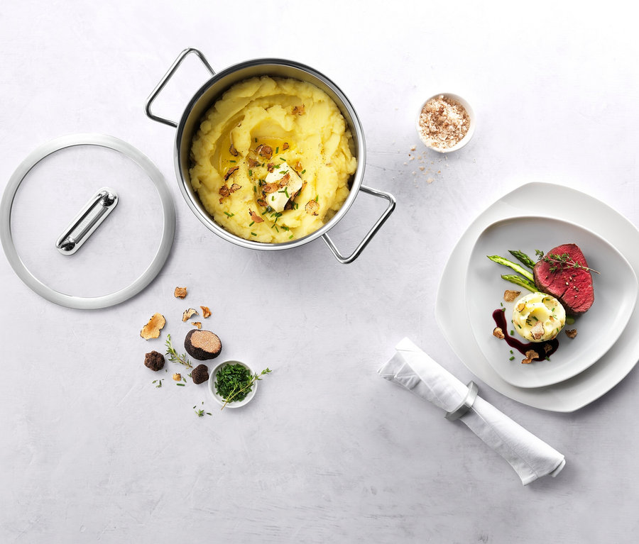 Ein Topf Kartoffelpüree mit Butter, Schnittlauch und Trüffelspänen, daneben ein Glasdeckel. Rechts ein Teller mit Rindfleisch, Spargel, Kartoffelgratin und Sauce. Daneben Trüffel, Kräuter und eine Serviette.