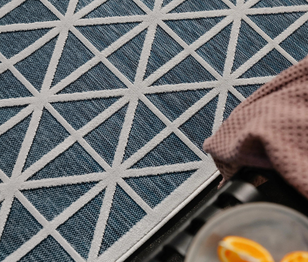 Blauer Teppich mit weißem geometrischem Dreiecksmuster, daneben ein rosafarbenes Tuch und unscharfe Orangenscheiben auf einem Teller.