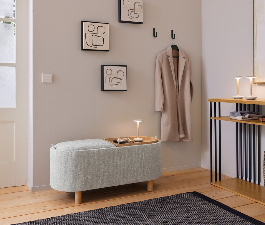 Ein gemütlicher Eingangsbereich mit einer hellgrauen Polsterbank, auf der ein Holztablett mit einer kleinen Lampe und Schlüsseln steht. Darüber hängen drei abstrakte Linienbilder und ein beiger Mantel an schwarzen Haken. Rechts davon steht eine Holzkonsole mit schwarzen Stützen, auf der zwei kleine Lampen, gefaltete Kleidung, ein Gürtel und eine geflochtene Tasche liegen. Ein dunkler gemusterter Teppich liegt auf dem Holzboden.