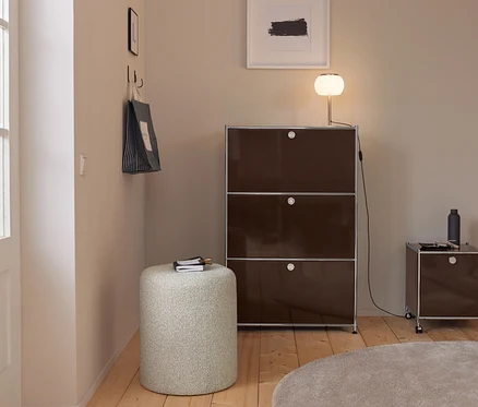 Moderner Eingangsbereich mit hellen Wänden, Holzboden und einem runden beigen Teppich. Ein hoher, dunkelbrauner Modulschrank mit einer leuchtenden Lampe steht neben einem kleineren Rollschrank. Ein heller zylindrischer Hocker mit Notizbuch ist im Vordergrund. Eine gestreifte Einkaufstasche hängt an Haken an der linken Wand.