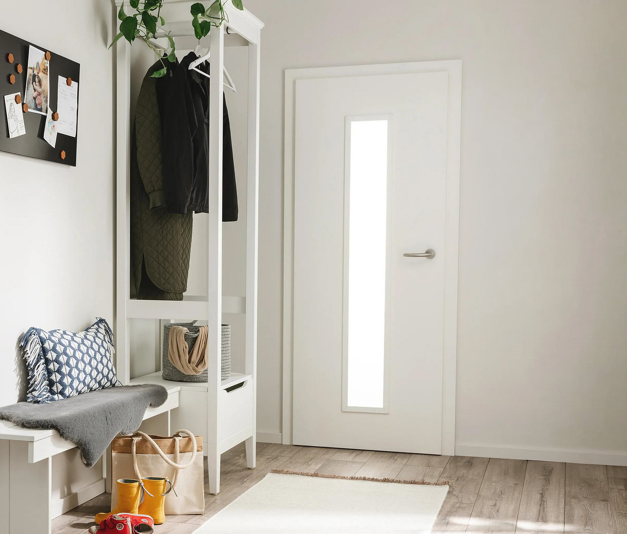 Flur mit weißer Garderobe und Bank links, einer weißen Tür rechts und hellem Holzboden. Die Garderobe trägt dunkle Kleidung und eine Pflanze, darunter ein grauer Korb. Die Bank hat ein blau gemustertes Kissen und einen grauen Überwurf. Auf dem Boden liegen gelbe Gummistiefel, eine Tragetasche und ein rotes Spielzeugauto. Eine schwarze Magnettafel hängt an der Wand. Ein hellbrauner benuta Läufer »Mia«, ca. 70 x 200 cm, liegt auf dem Boden.
