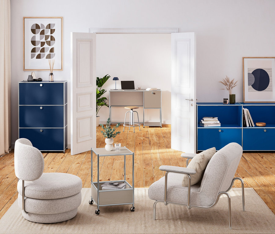 Modernes Wohnzimmer und Homeoffice. Im Vordergrund stehen hellgraue Polstersessel und ein Rollbeistelltisch auf einem Teppich, flankiert von dunkelblauen Modulschränken. Durch eine offene Tür ist ein Homeoffice mit einem hellgrauen Schreibtisch und einem Hocker mit Holzsitzfläche sichtbar.
