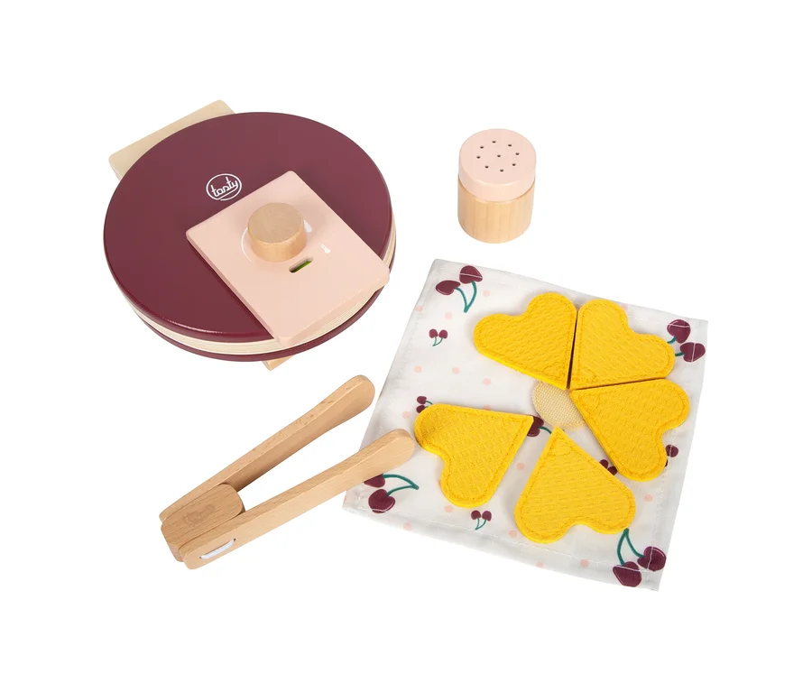 Ein rotes small foot Waffeleisen-Set »tasty« mit einem Holzstreuer, einer Holzzange und vier gelben herzförmigen Spielwaffeln auf einer Kirschmuster-Serviette.