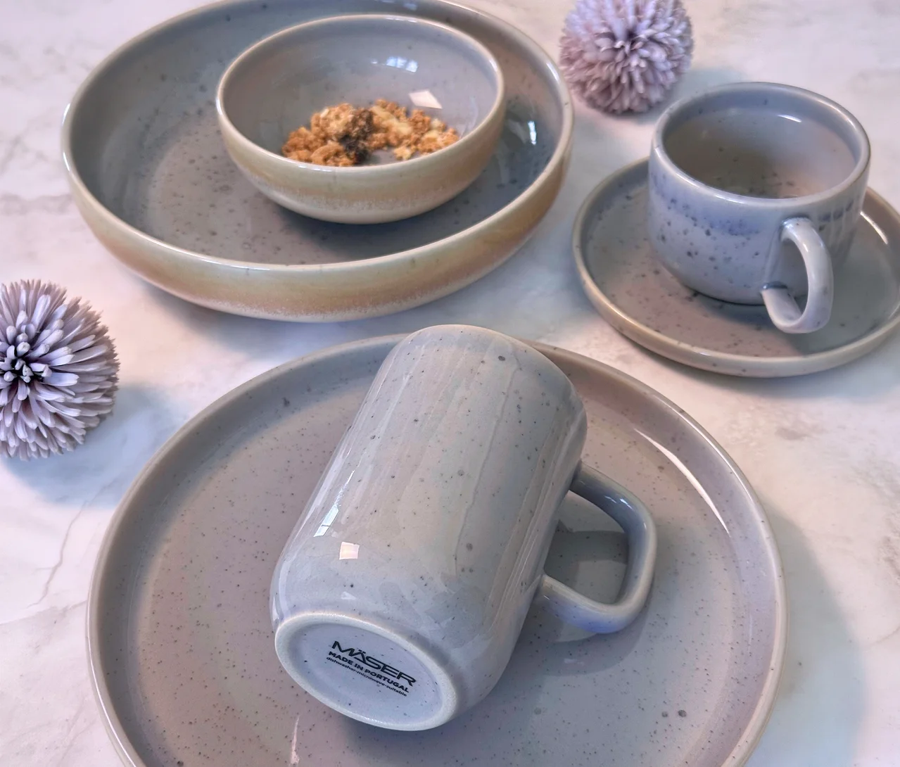 Mäser Tafelservice »Candy« und Frühstücks-Set »Candy«: Eine Anordnung aus grauen, gesprenkelten Keramikschalen, Tassen und Tellern auf einer Marmoroberfläche.